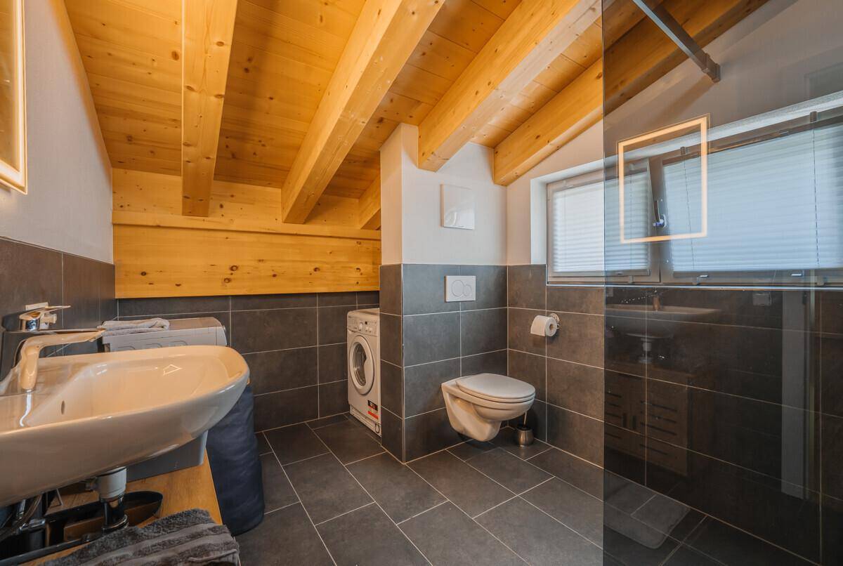 Modernes Badezimmer im Dachgeschoss mit Holzbalken, dunklen Fliesen und Waschmaschine.