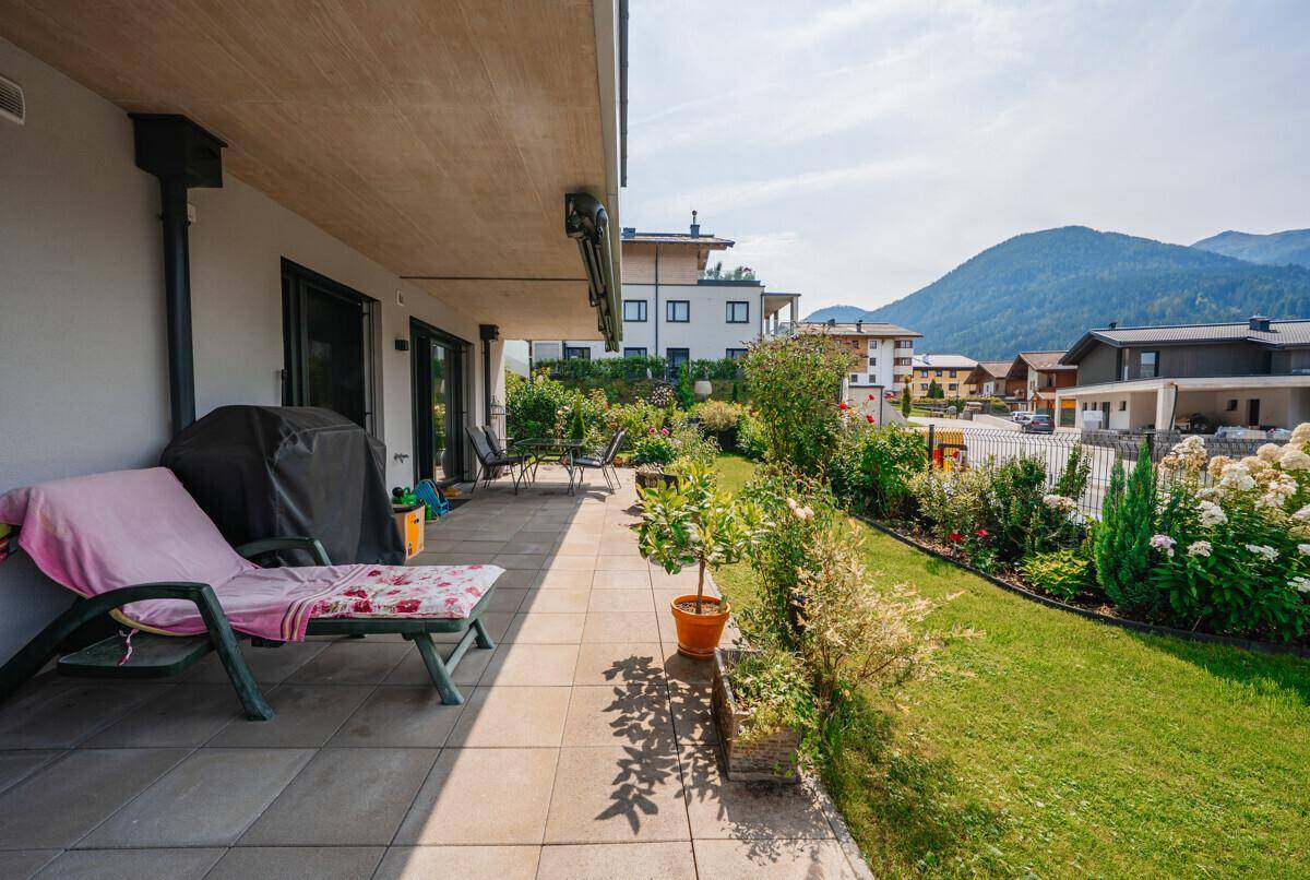 Sonniger Terrassenbereich mit Liegestuhl, Blumenbeeten und Zugang zum Garten.
