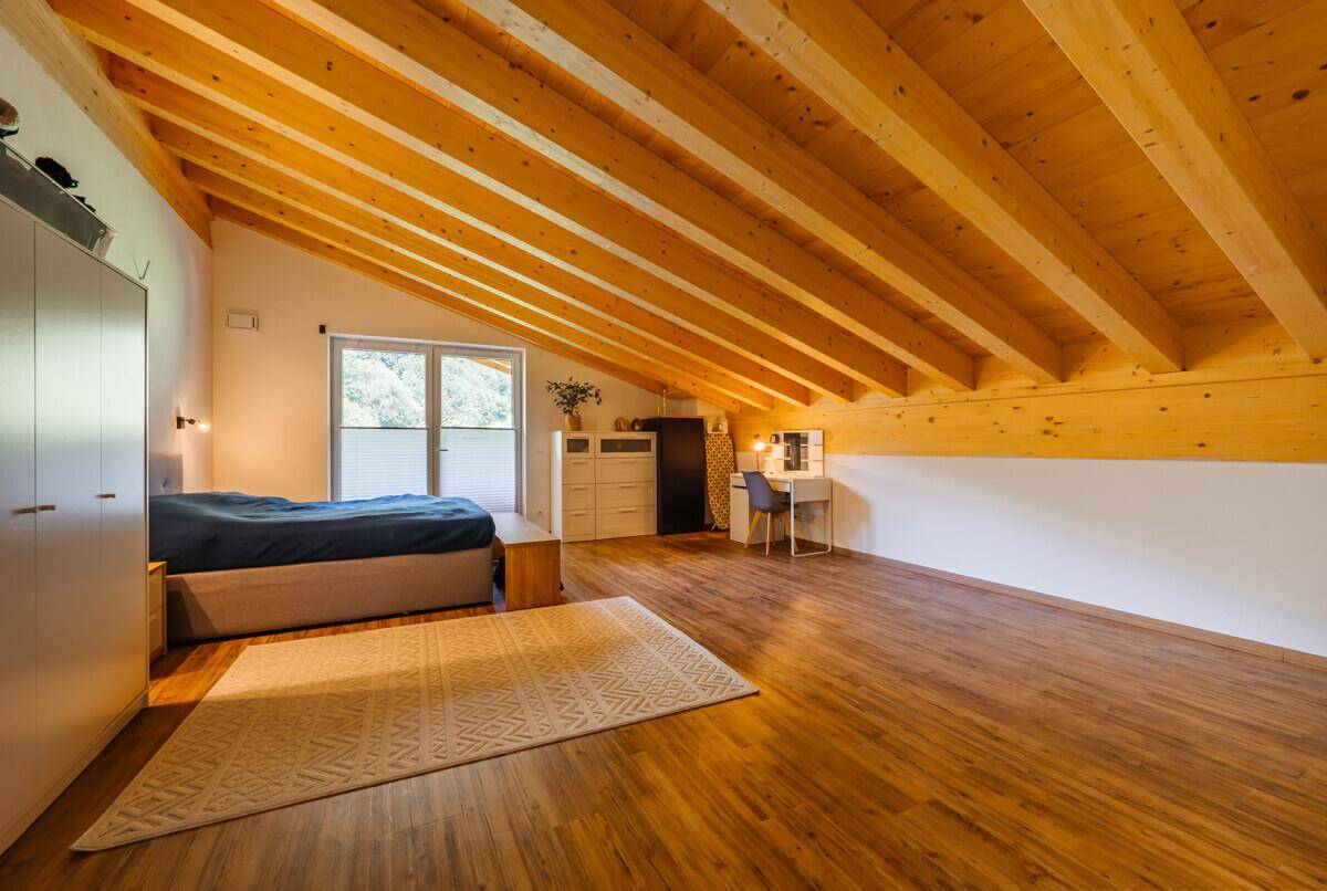 Helles Schlafzimmer im Dachgeschoss mit sichtbaren Holzbalken, Holzboden und Blick ins Grüne.