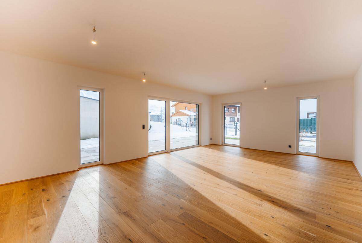 Heller, leerer Wohnraum mit Holzboden und großen Fenstern, die viel Tageslicht hereinlassen.