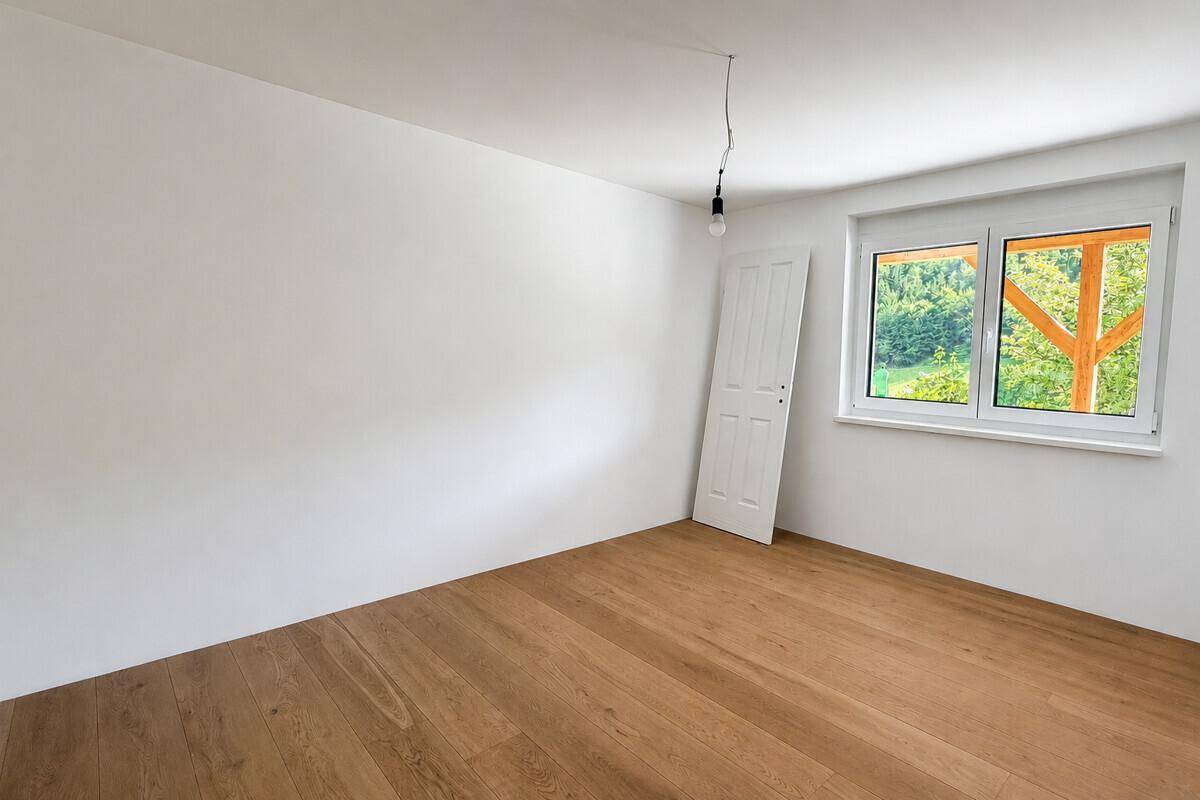 Geräumiges, leeres Zimmer mit Holzboden und Fenster, bietet einen schönen Blick ins Grüne.
