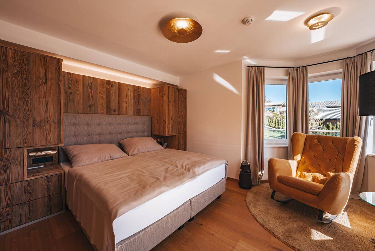 Schlafzimmer mit Doppelbett, Holzwand, Sessel und Fenster mit Blick ins Grüne.
