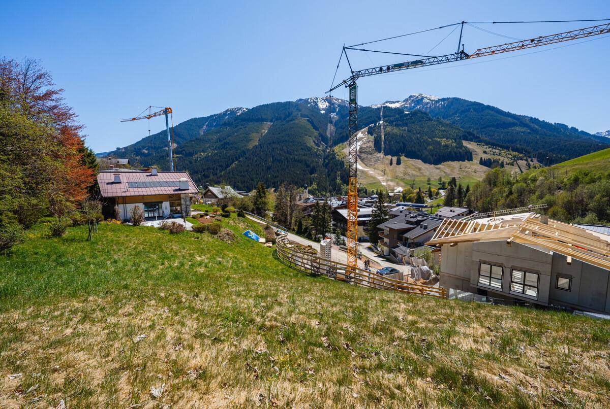 Weitläufige grüne Landschaft mit einer Baustelle, bestehenden Häusern und majestätischen Bergen im Hintergrund.