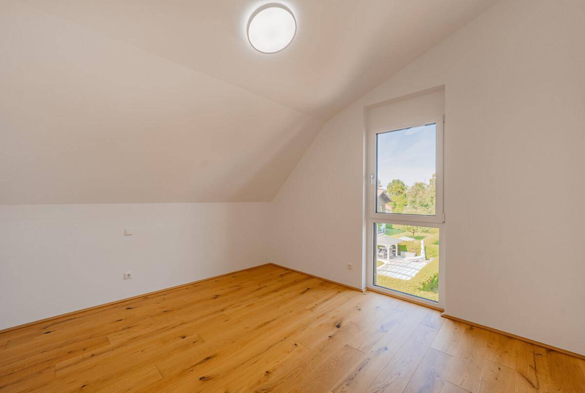 Helles Zimmer mit Holzboden, Dachschrägen und einem hohen Fenster mit Blick ins Grüne.