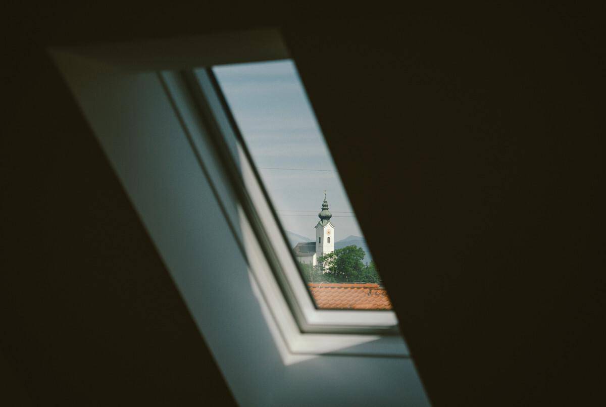 Blick durch ein Dachfenster auf einen Kirchturm in der Ferne, umgeben von Grün.