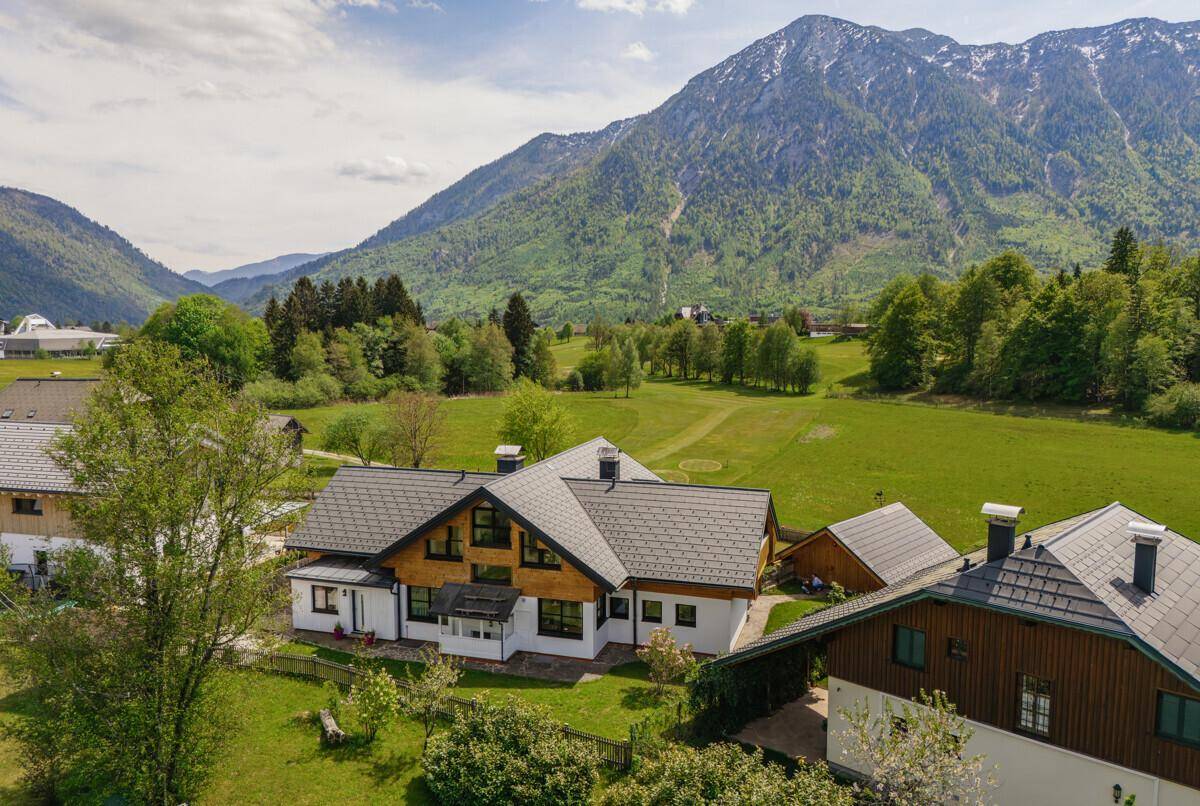 Haus in alpiner Landschaft mit Bergblick und grünen Wiesen unter blauem Himmel.