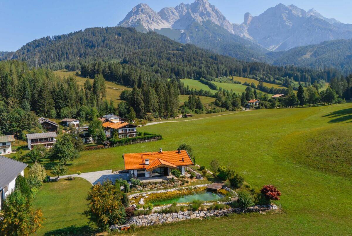 Luftaufnahme des Hauses mit Garten, Teich und umliegender Naturlandschaft mit Bergen.