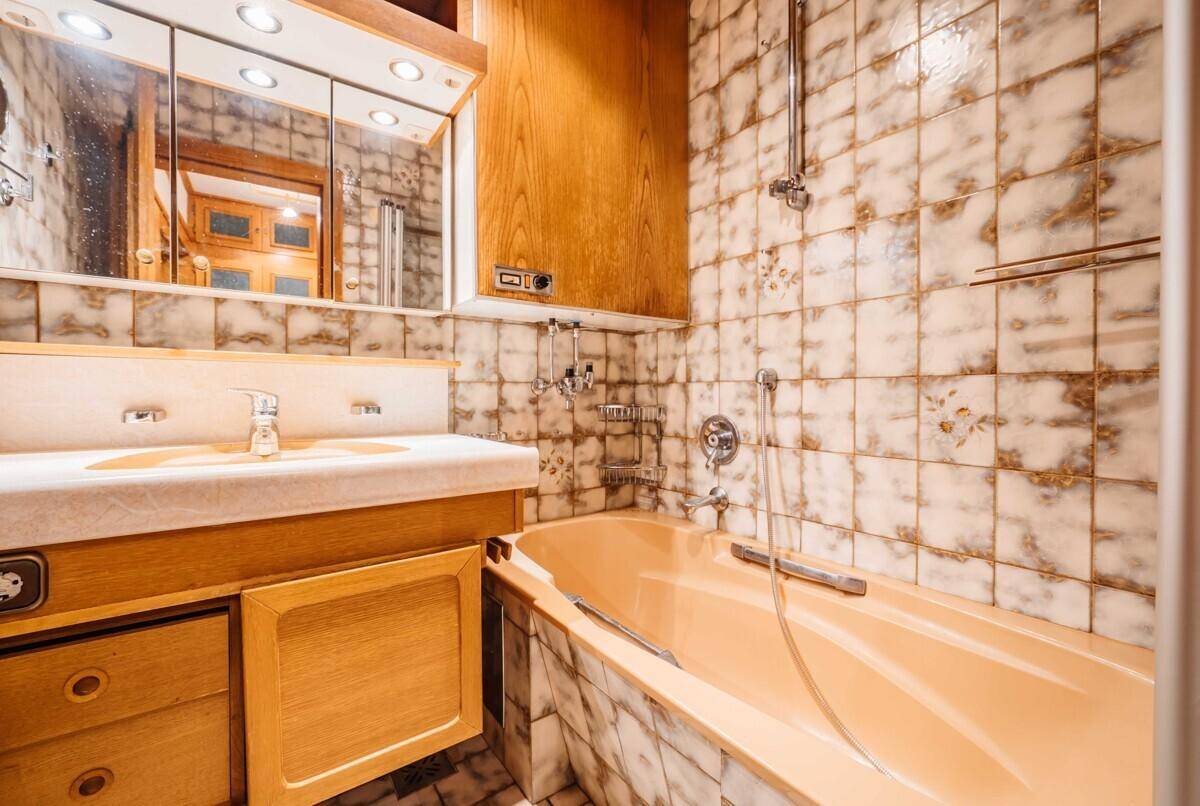 Badezimmer im 80er-Jahre-Stil mit beigefarbenen Fliesen, Badewanne und Holzschrank unter dem Spiegel.