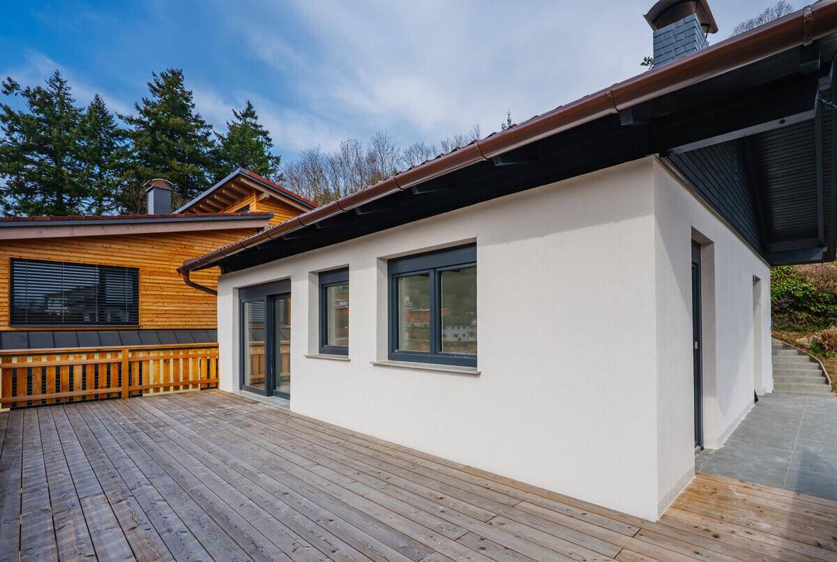Große Holzterrasse neben einem modernen Haus mit weißer Fassade und dunklen Fenstern.