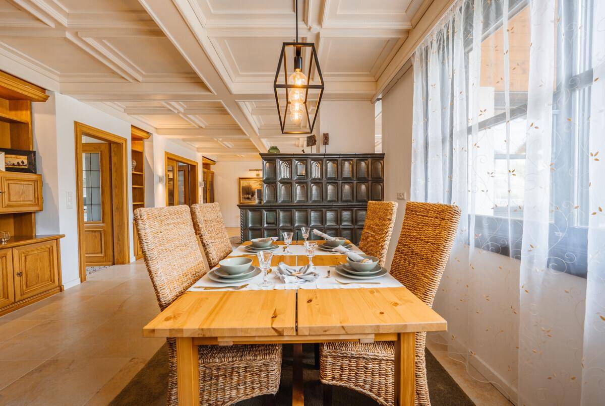 Einladender Essbereich mit Holztisch, Korbstühlen und traditionellem Kachelofen im Hintergrund.