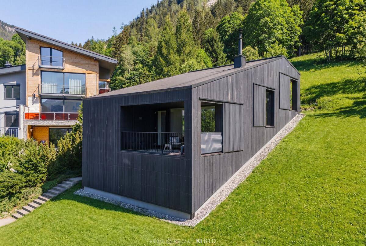 Zeitgenössisches Haus mit dunkler Holzverkleidung und Balkon, umgeben von grüner Natur und Bergen.