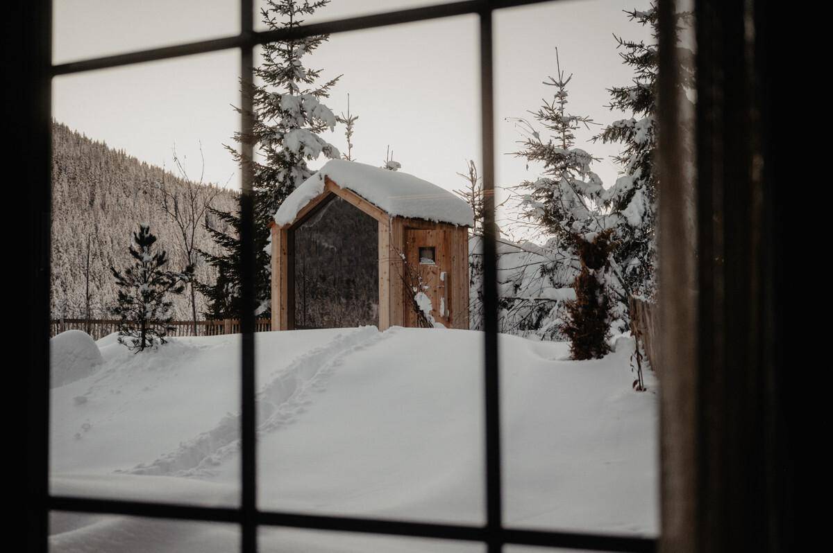 Blick aus dem Fenster auf eine verschneite Holzhütte in einer winterlichen Berglandschaft.