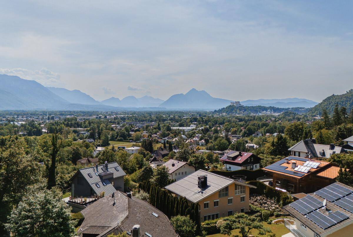 Panoramablick über eine Wohnsiedlung mit Dächern und Bergen im Hintergrund unter blauem Himmel.
