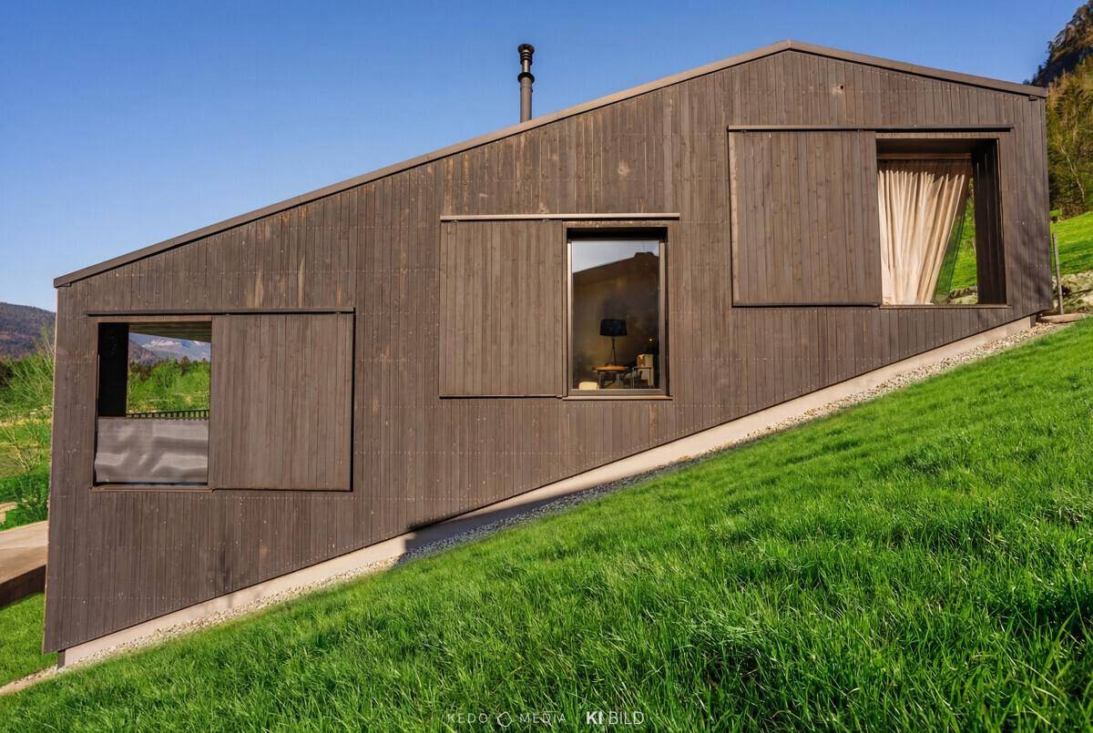 Schräge schwarze Holzfassade des modernen Hauses, eingebettet in eine grüne Hügellandschaft mit Fenstern.