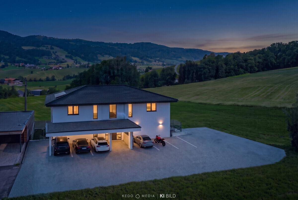 Modernes Wohngebäude mit beleuchtetem Carport und Parkplätzen bei Einbruch der Dunkelheit.