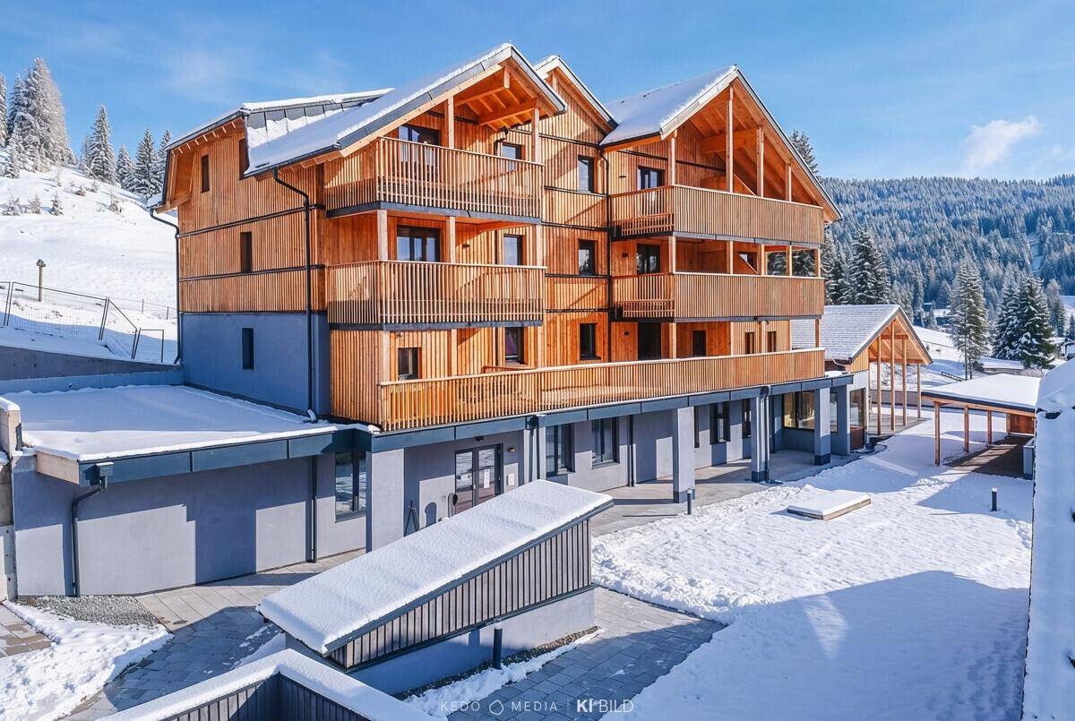 Moderne Chalet-Anlage im Winter mit schneebedeckten Dächern und Balkonen vor einer Waldkulisse.