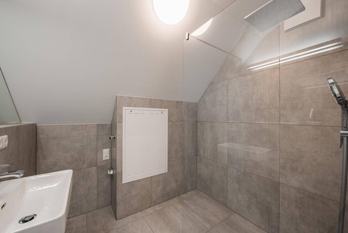Modernes Badezimmer mit Duschbereich, Waschbecken und Dachschräge, komplett gefliest in Grautönen.