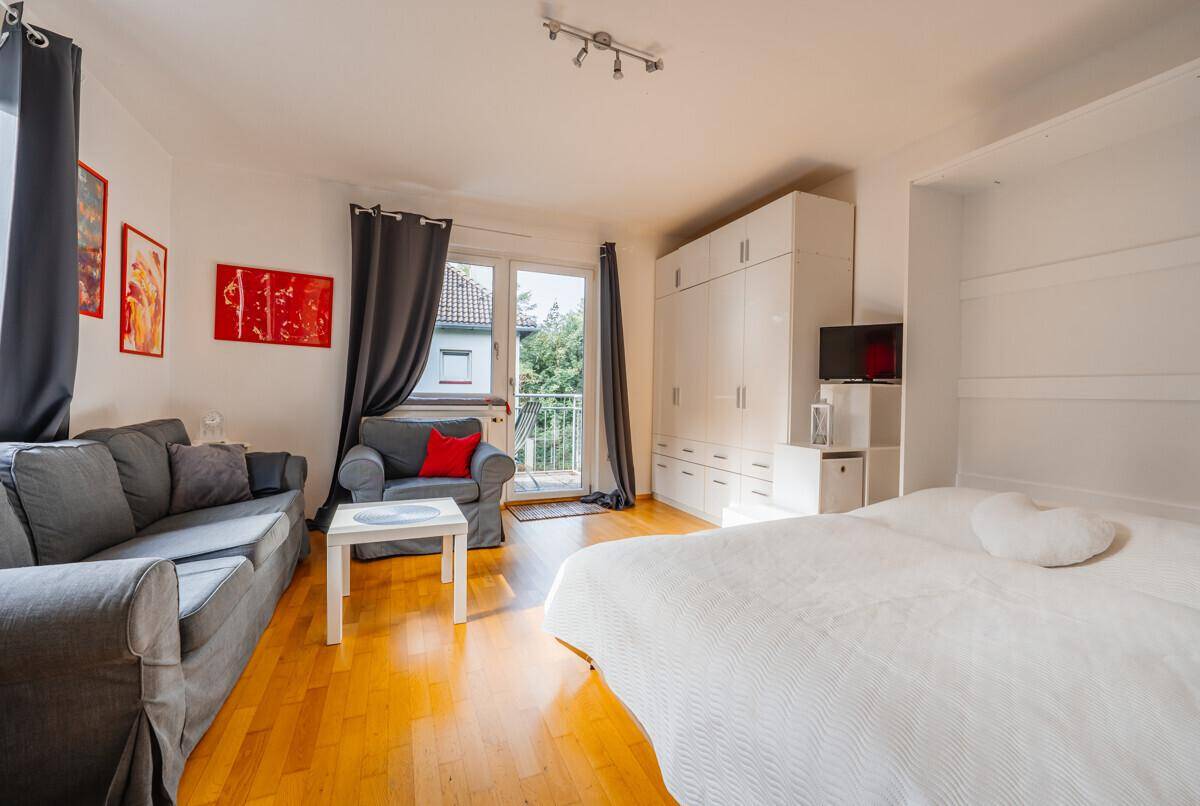 Heller Wohn- und Schlafbereich mit Holzboden, Sofa, Schrankwand und Zugang zum Balkon.
