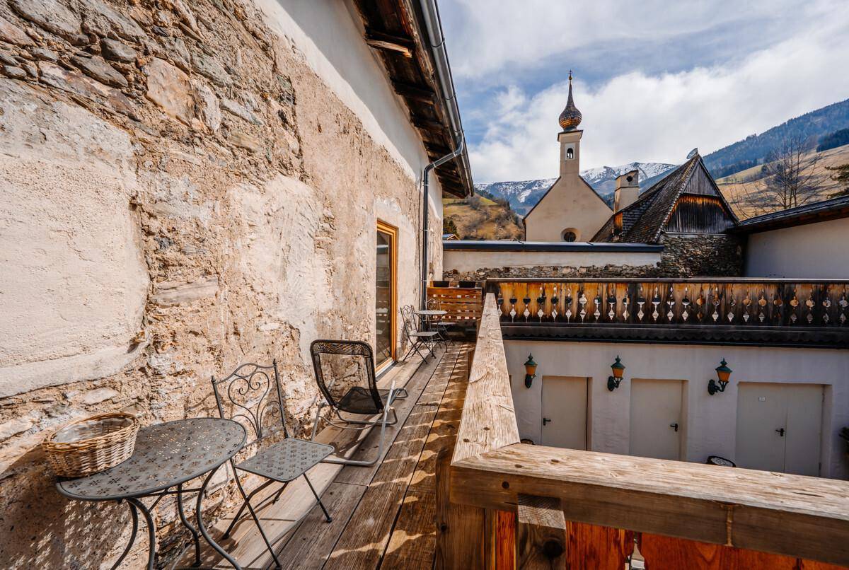 Balkon mit Holzboden, schmiedeeisernen Möbeln und Blick auf die Berge und Kirche.