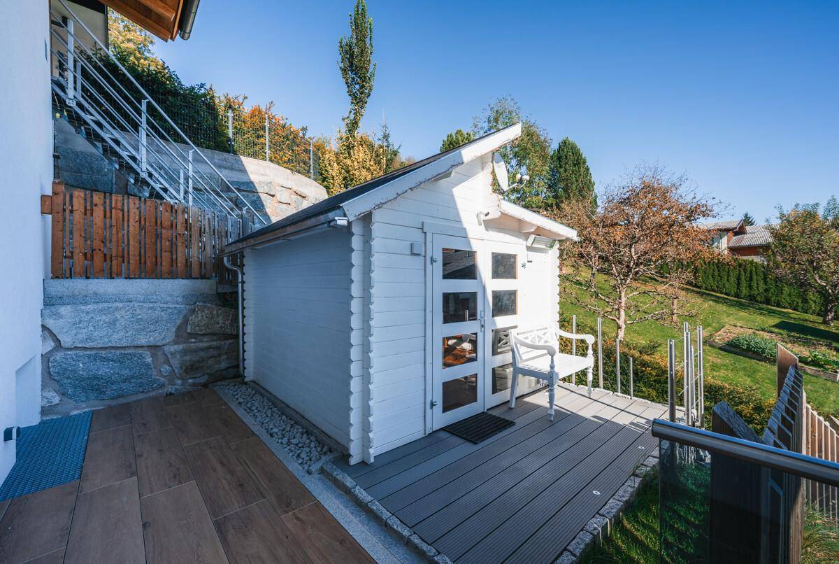 Kleines weißes Gartenhaus mit Terrasse und Sitzbank in sonniger Lage.