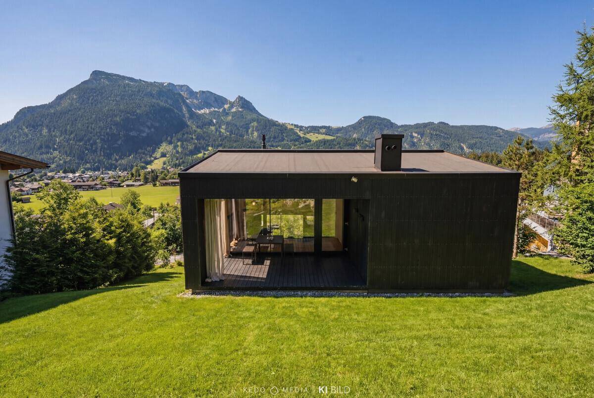 Modernes Haus mit Flachdach und großen Glasfronten, umgeben von grünen Wiesen und Bergen.