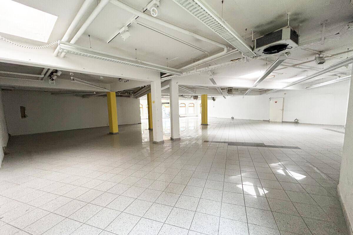Großer, leerer Gewerberaum mit Fliesenboden, gelben Säulen und sichtbaren Installationen an der Decke.