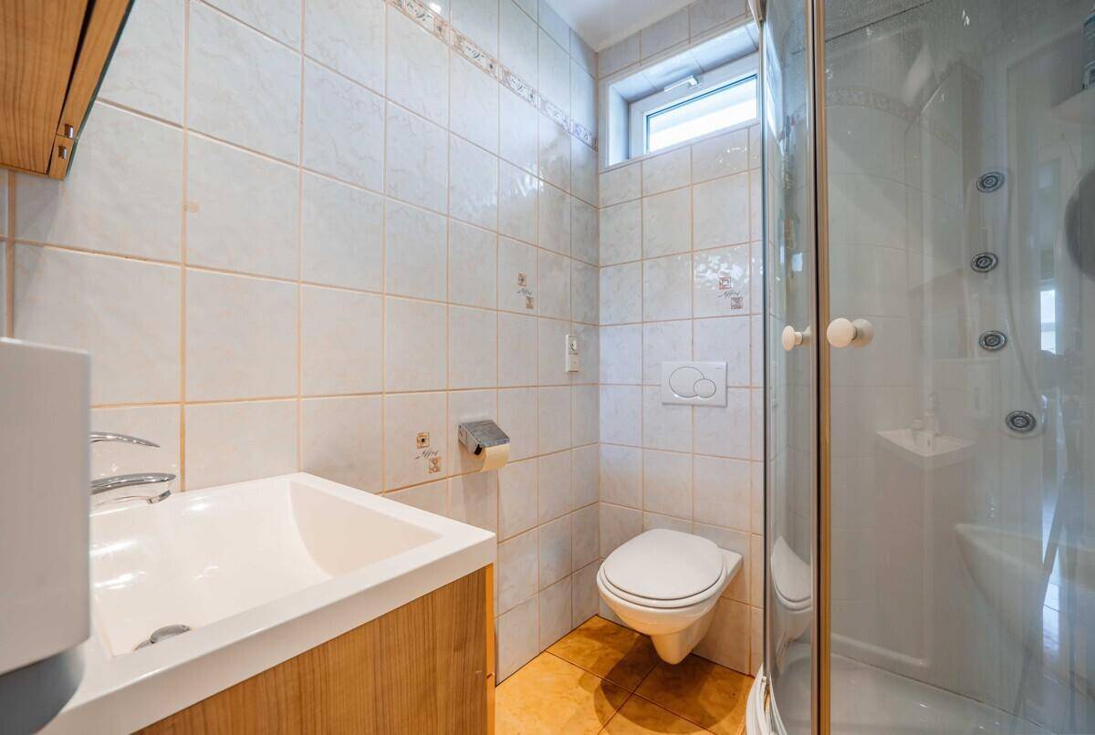 Badezimmer mit weißen Fliesen, Dusche, WC und einem kleinen Fenster für Tageslicht.