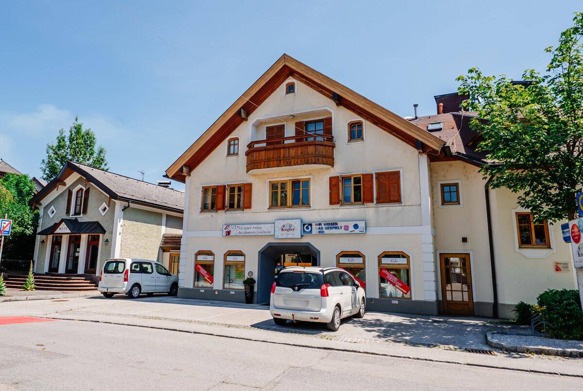 Traditionelles Gebäude mit Holzbalkon, Fensterläden und Geschäften im Erdgeschoss an einer Straße.