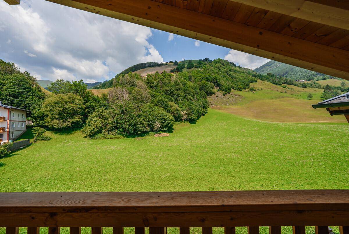 Weitläufiger Blick von einem Balkon auf grüne Wiesen und bewaldete Hügel unter blauem Himmel.