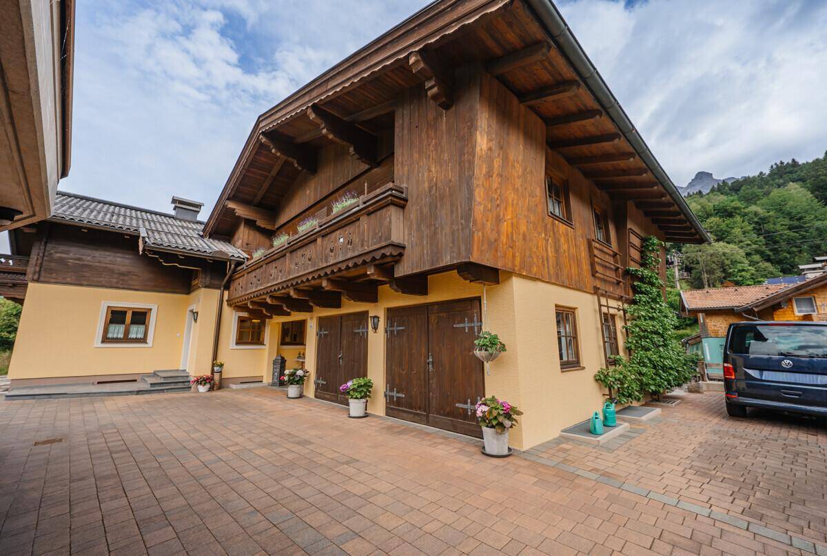 Traditionelles Haus mit Holzbalkon und gepflasterter Auffahrt, umgeben von grüner Natur.