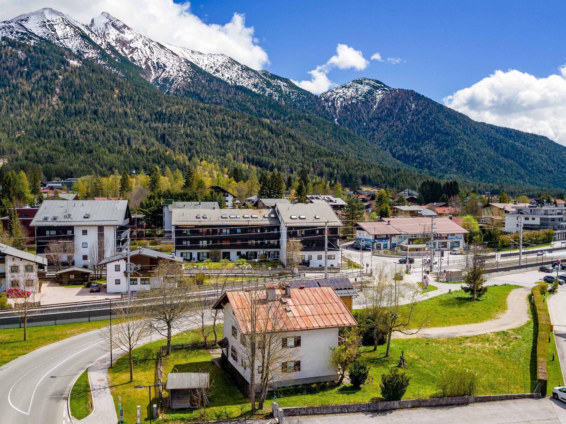 Weitläufige Ansicht eines alpinen Dorfes, umgeben von bewaldeten Bergen und grünen Wiesen.