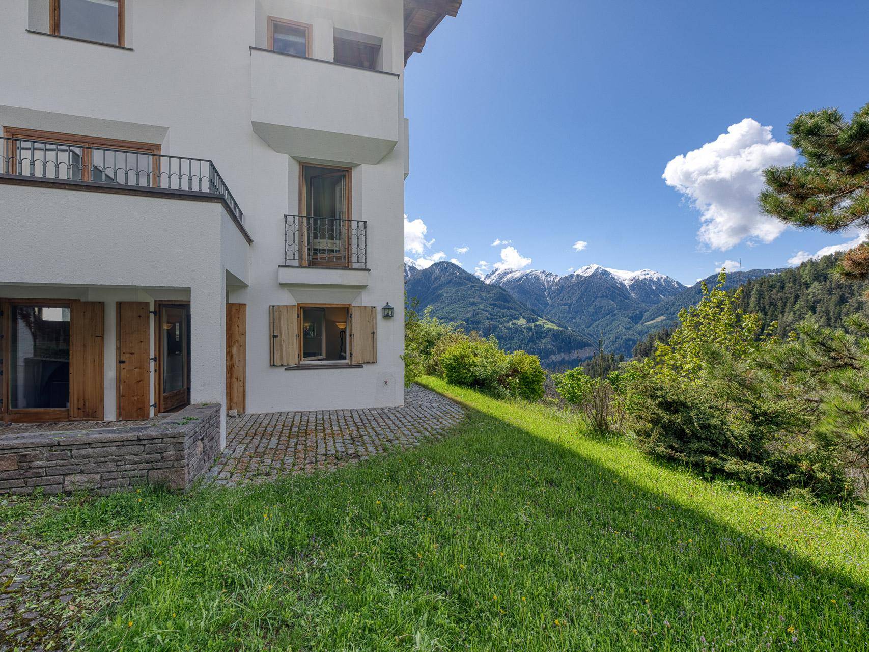 Grüne Rasenfläche neben dem Haus mit gepflasterter Terrasse und weitem Bergblick.