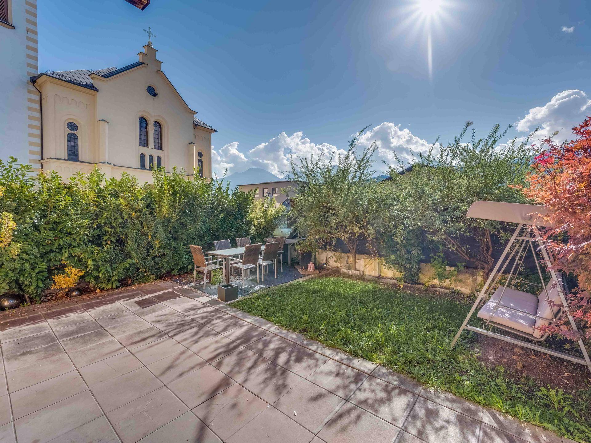 Sonniger Gartenbereich mit gepflasterter Terrasse, Essbereich und Schaukel vor historischer Kulisse.