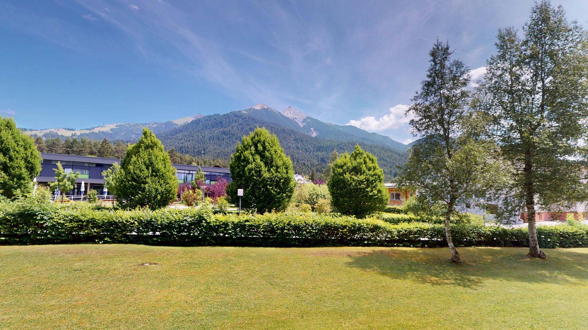 Weitläufige Grünfläche mit Bäumen und einem beeindruckenden Bergpanorama im Hintergrund.