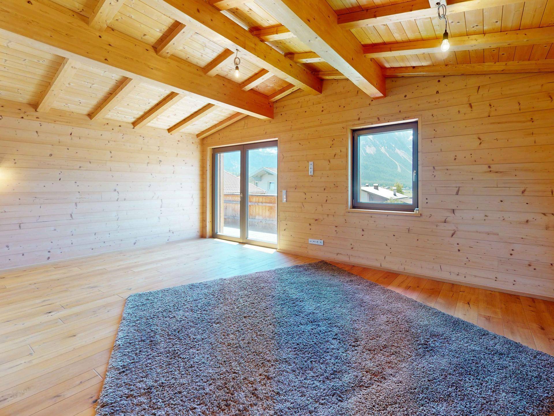 Geräumiger, holzvertäfelter Raum mit sichtbaren Deckenbalken und Zugang zu einem Balkon.