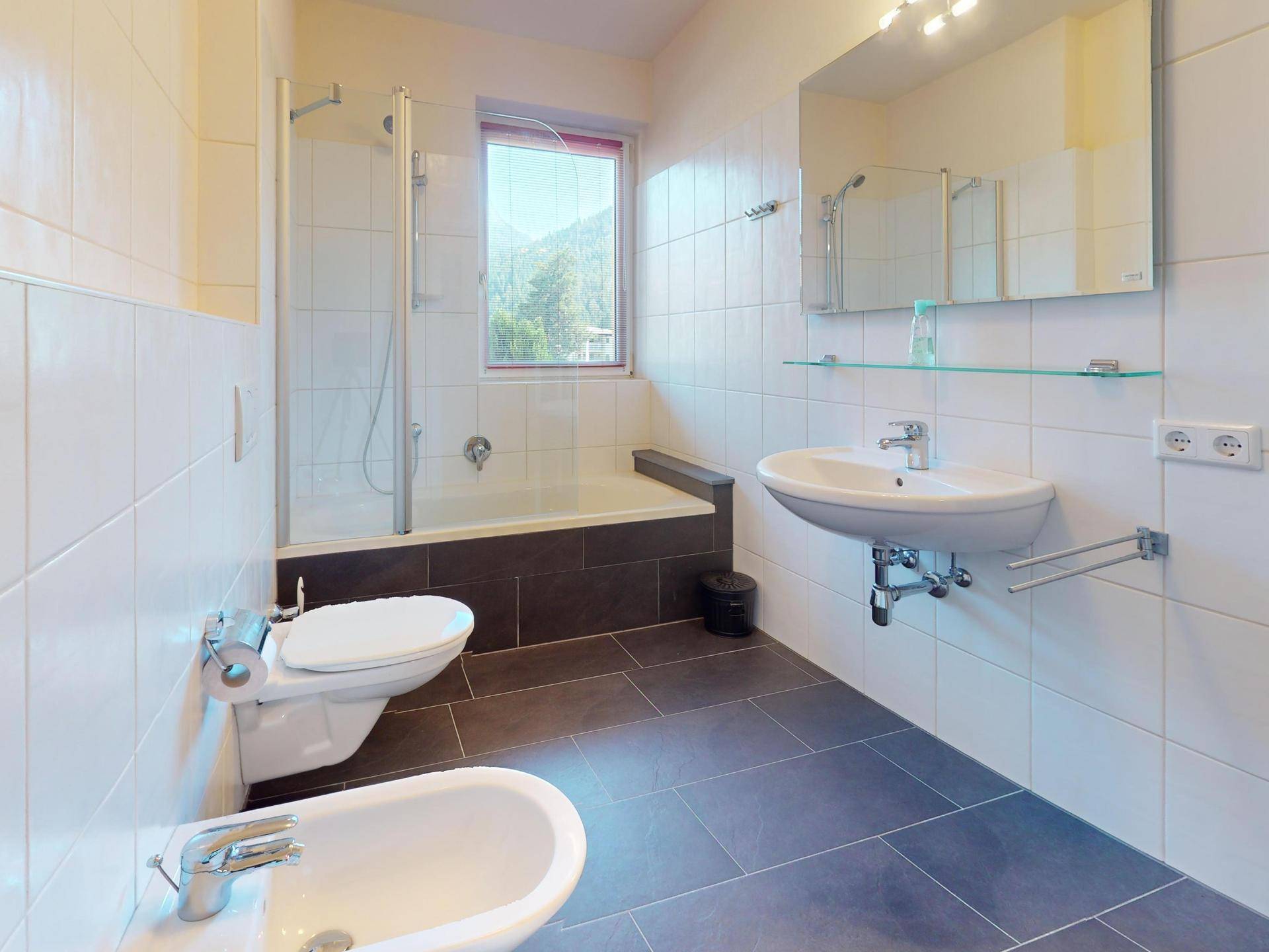 Modernes Badezimmer mit weißen Fliesen, Dusche, Badewanne, Bidet und Fenster.