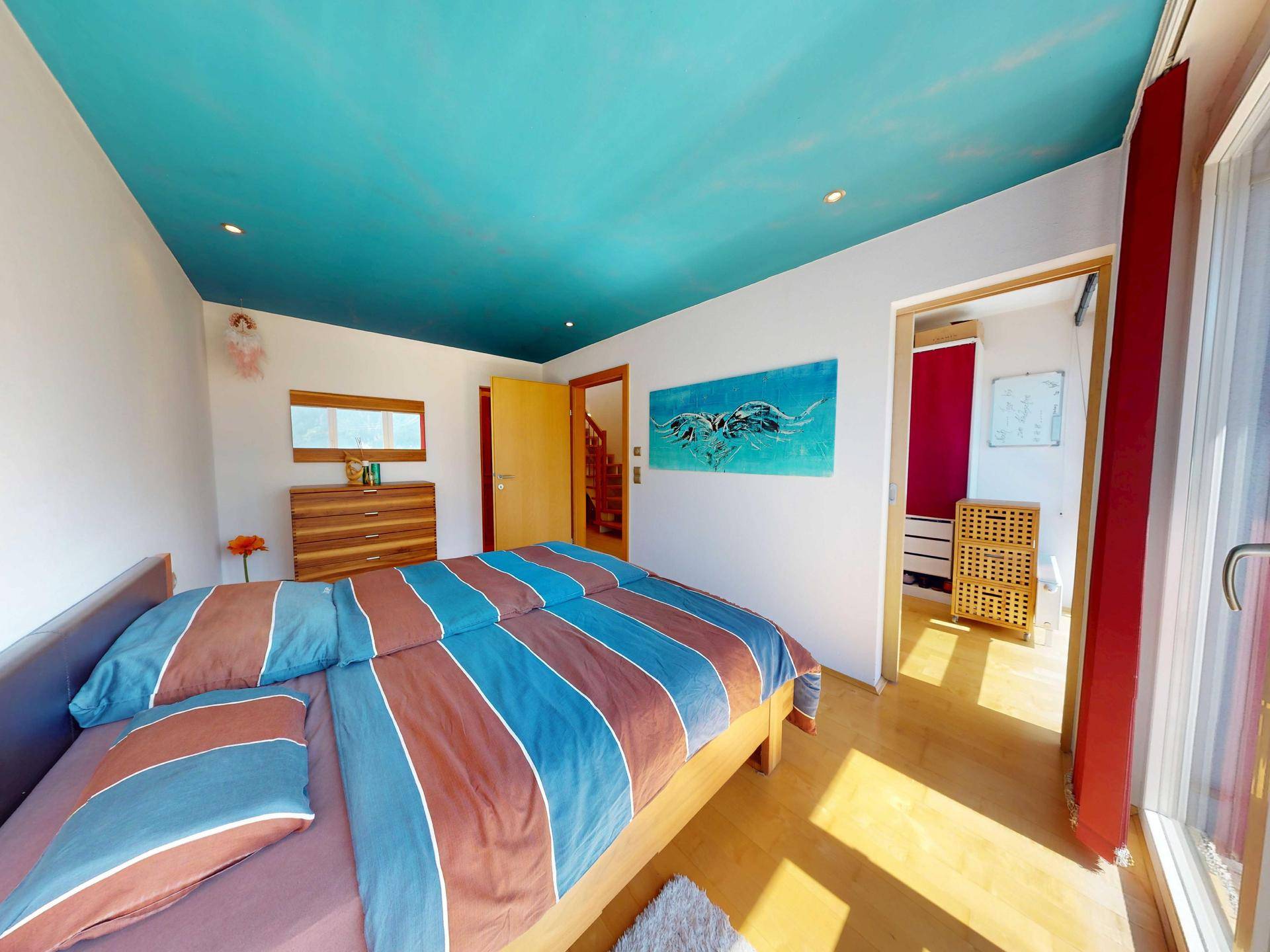 Gemütliches Schlafzimmer mit farbiger Decke, Holzbett und viel natürlichem Licht durch das Fenster.