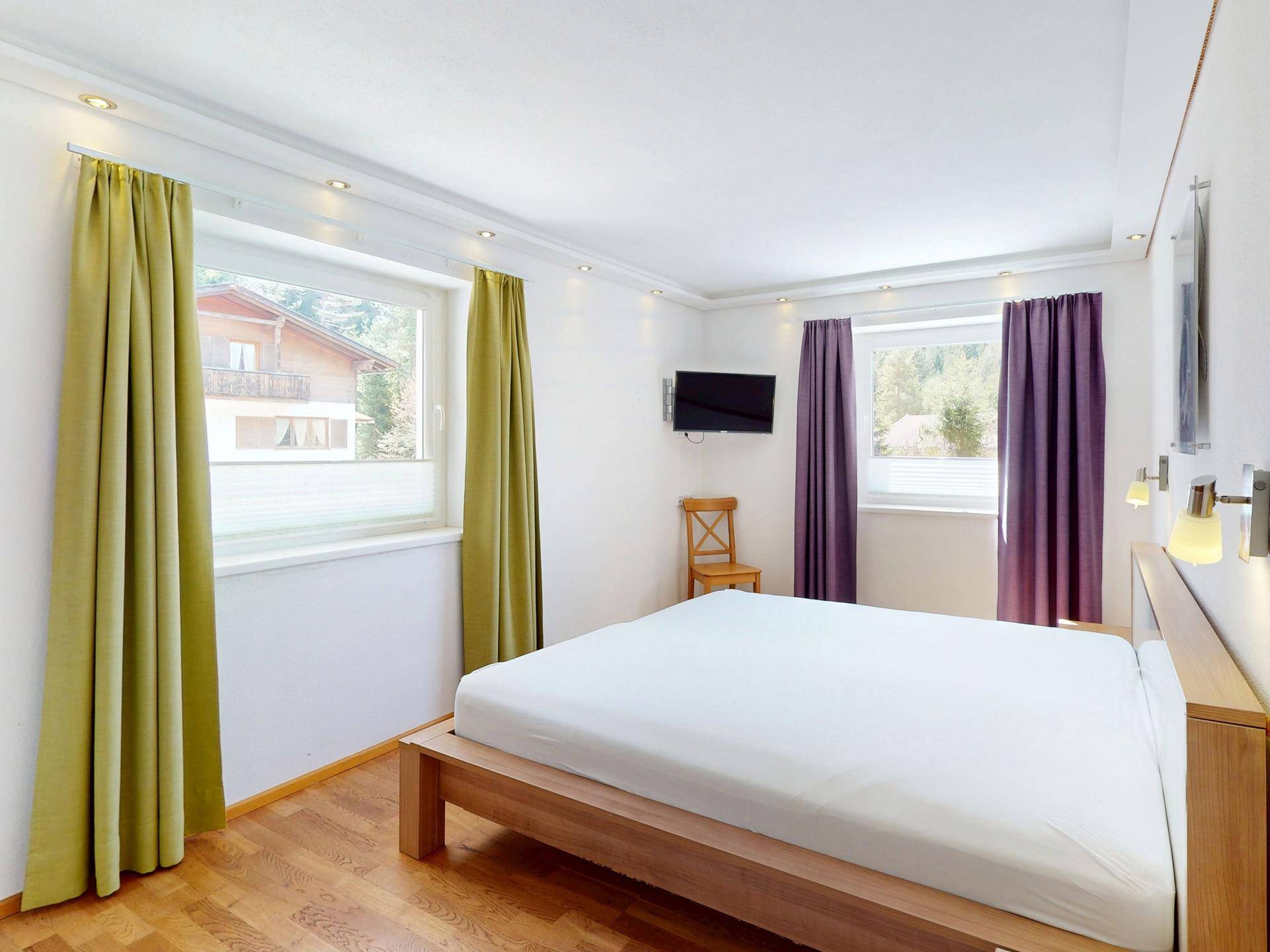 Gemütliches Schlafzimmer mit Holzbett, Fernseher und zwei Fenstern für viel Tageslicht.