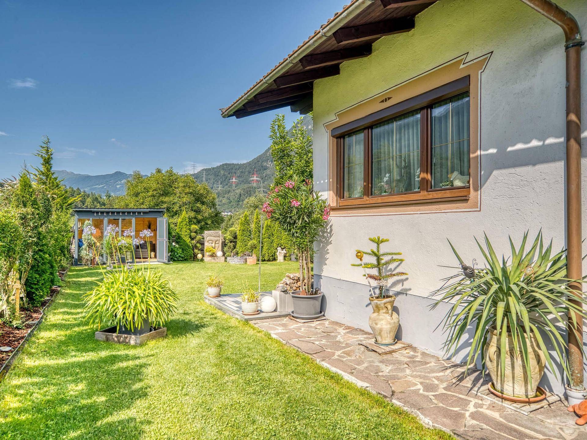 Sonnige Gartenseite des Hauses mit Rasenfläche, Pflanzen und einem Gartenhaus im Hintergrund.
