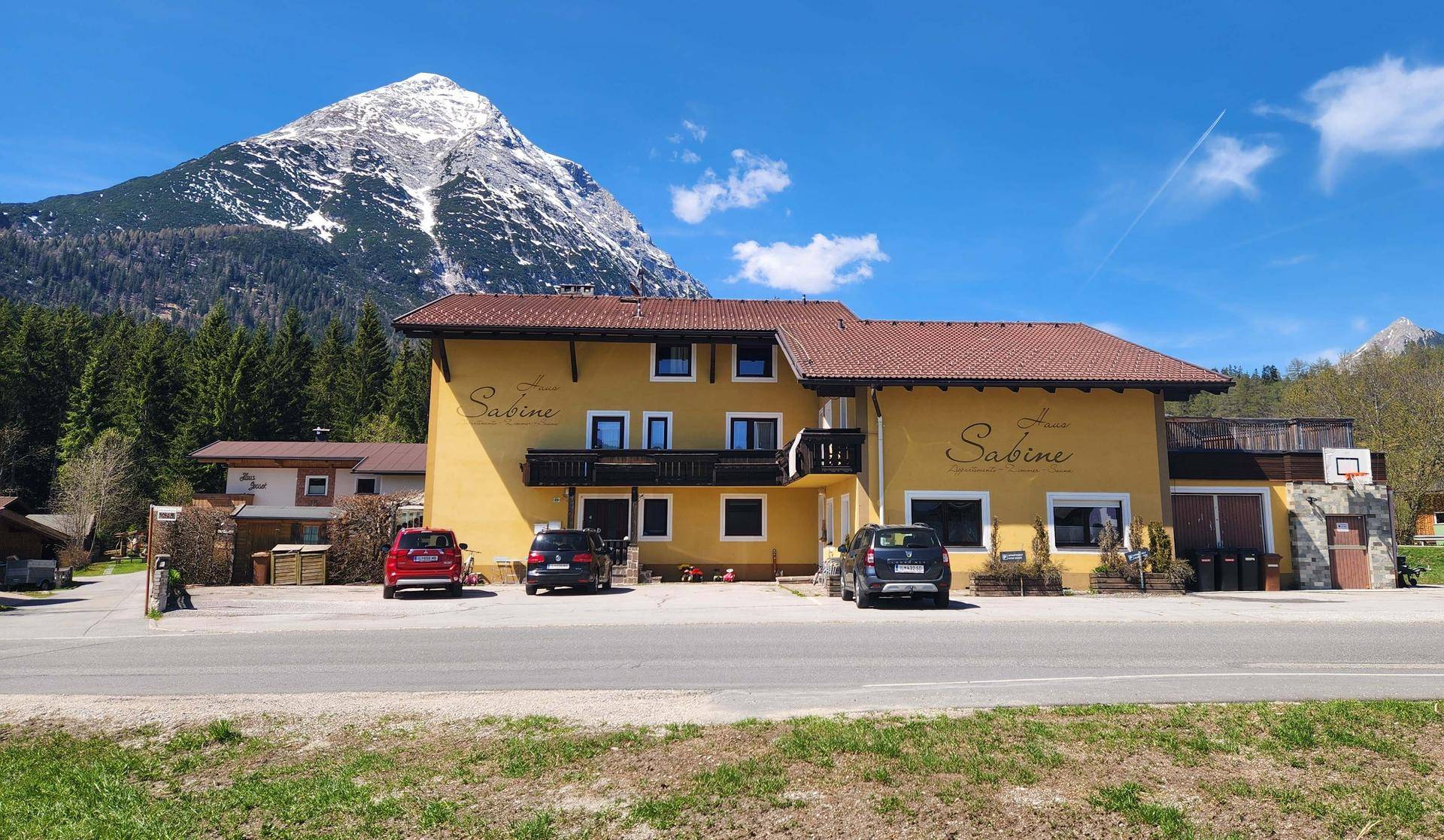 Gelbes Gebäude mit rotem Dach und beeindruckendem Bergblick unter blauem Himmel.