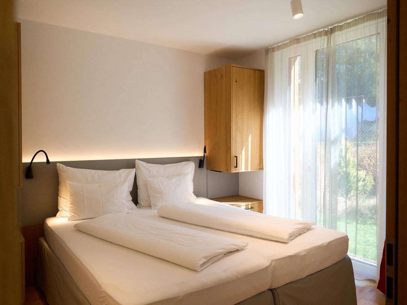 Helles Schlafzimmer mit Doppelbett, Holzelementen und großem Fenster mit Blick ins Grüne.