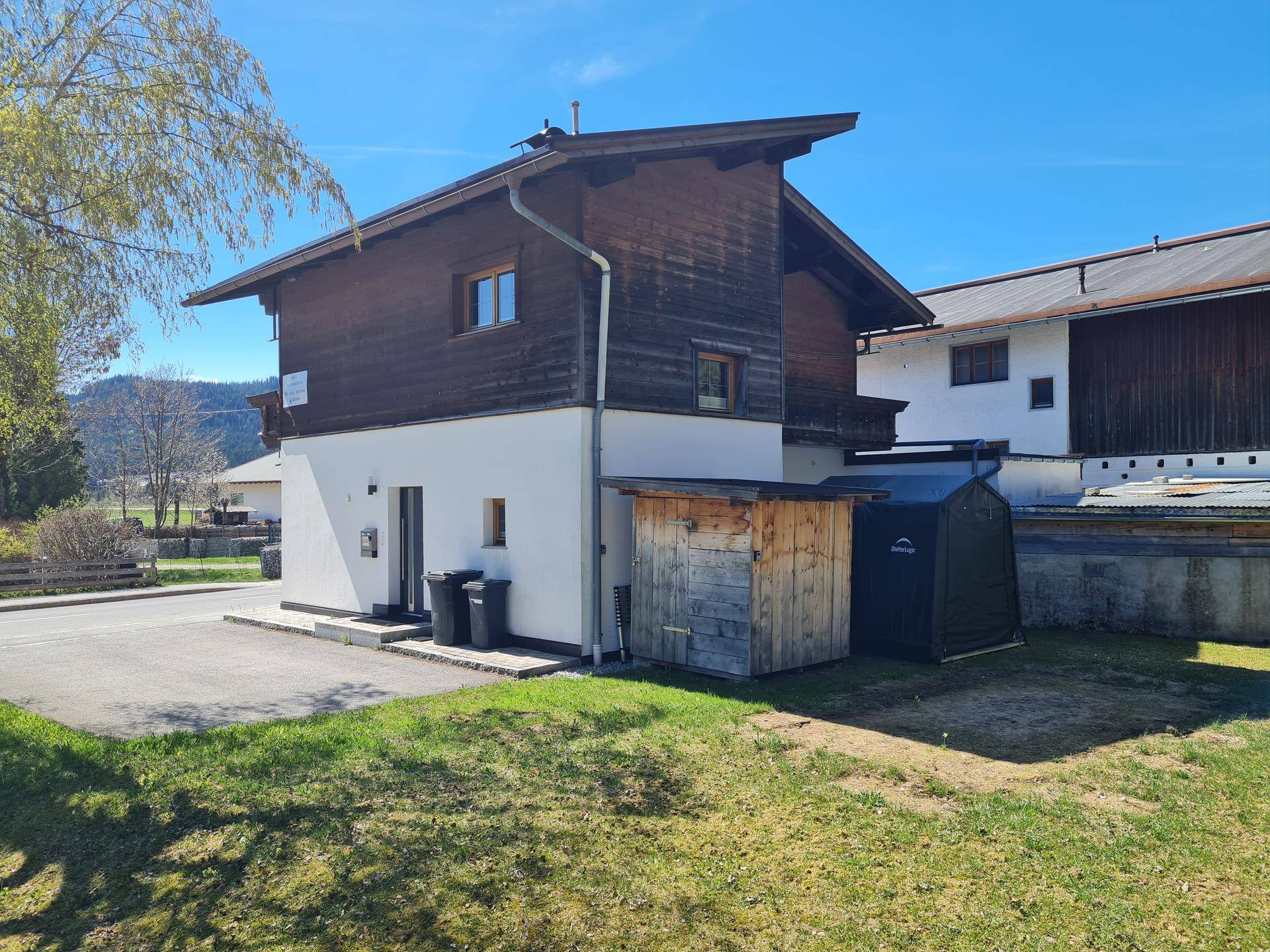 Traditionelles Haus mit Holzfassade und weißem Sockel, umgeben von Grünflächen und Bäumen.