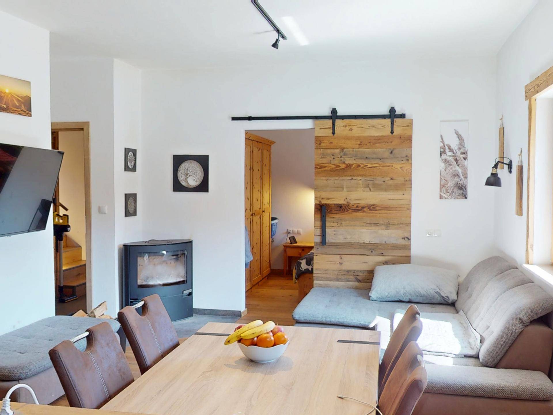 Heller Wohn- und Essbereich mit Holzboden, Kaminofen und rustikaler Schiebetür zum Schlafzimmer.