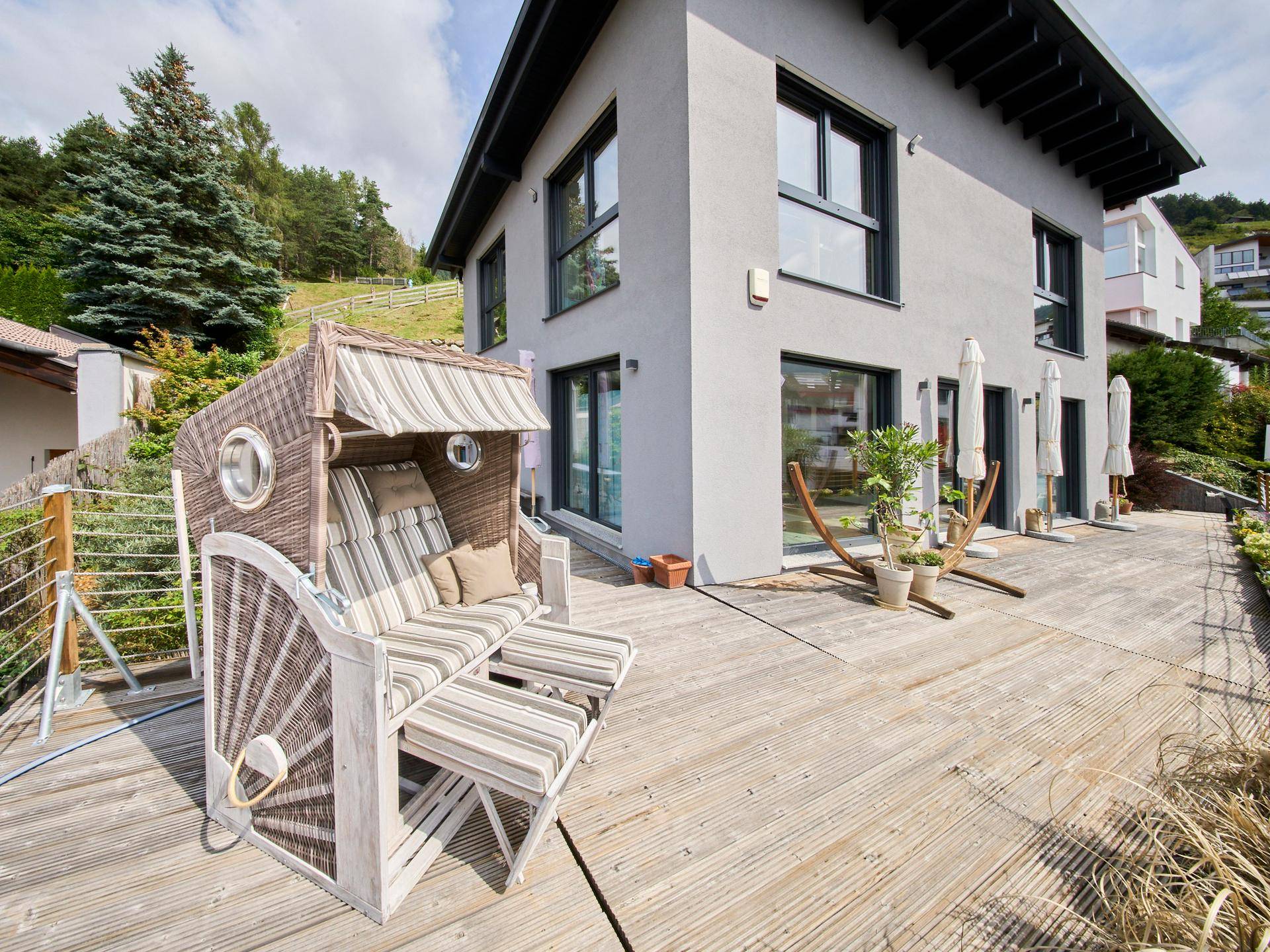 Große Holzterrasse mit Strandkorb und Blick auf das moderne Haus und die grüne Umgebung.
