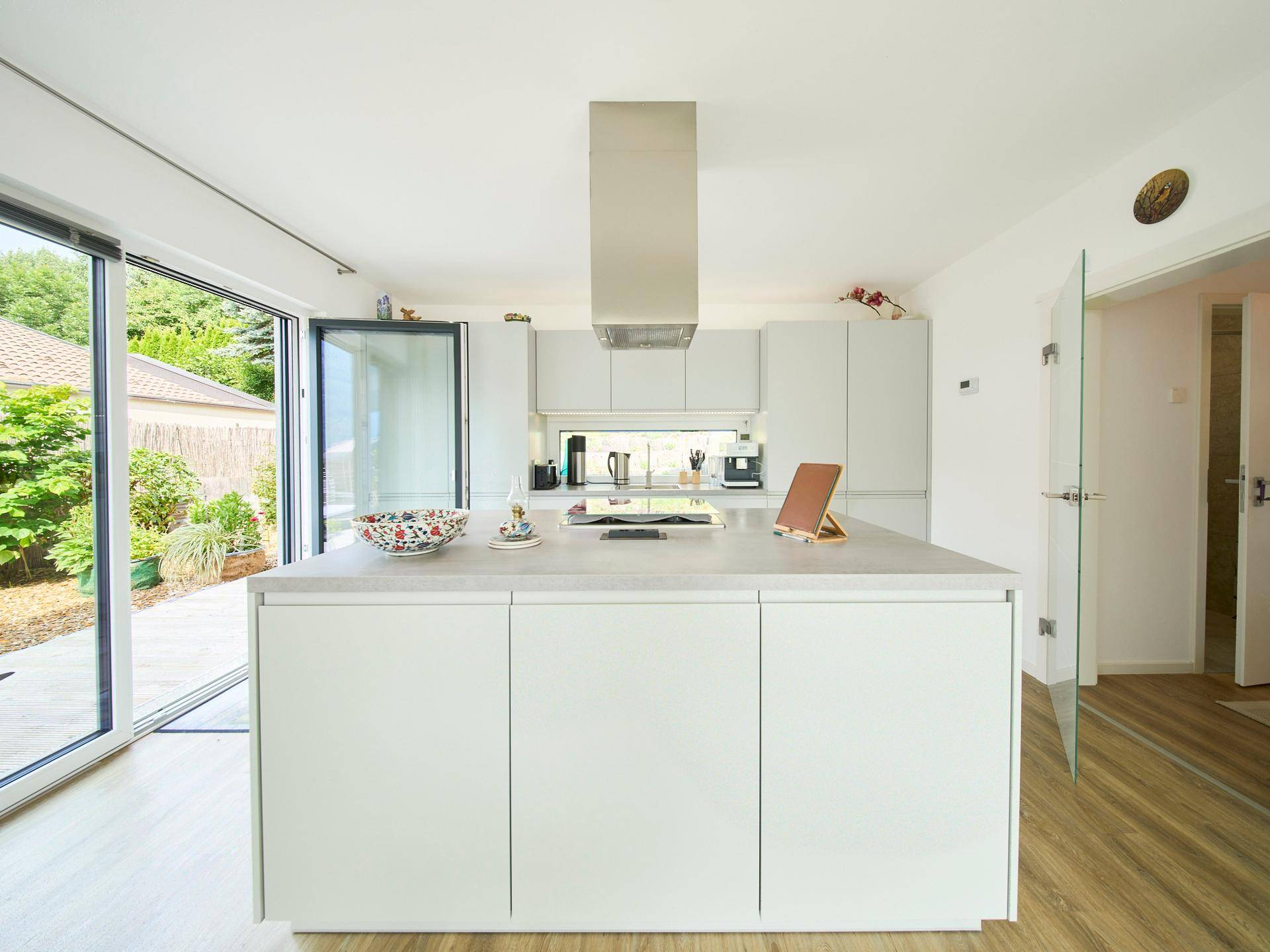 Moderne weiße Küche mit Kochinsel, Dunstabzugshaube und Zugang zur Terrasse.