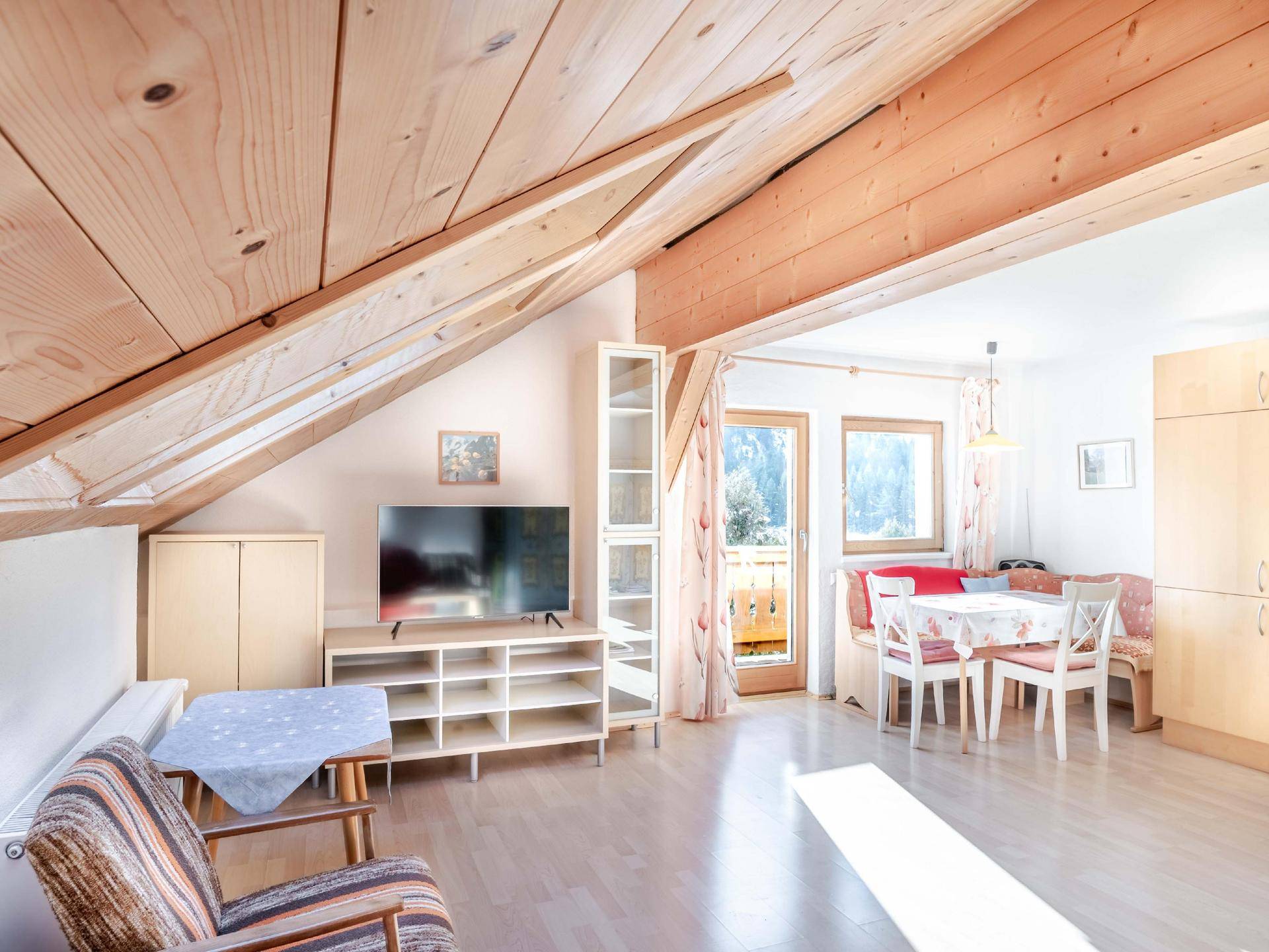 Gemütlicher Wohn- und Essbereich im Dachgeschoss mit Holzbalken, Fernseher und Zugang zum Balkon.