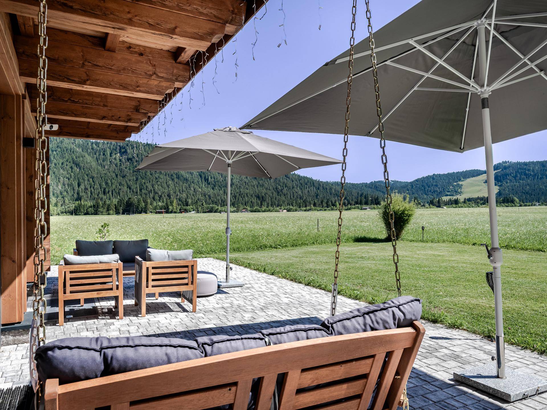 Gepflasterte Terrasse mit bequemen Sitzgelegenheiten, Sonnenschirmen und einer Schaukelbank mit Blick ins Grüne.