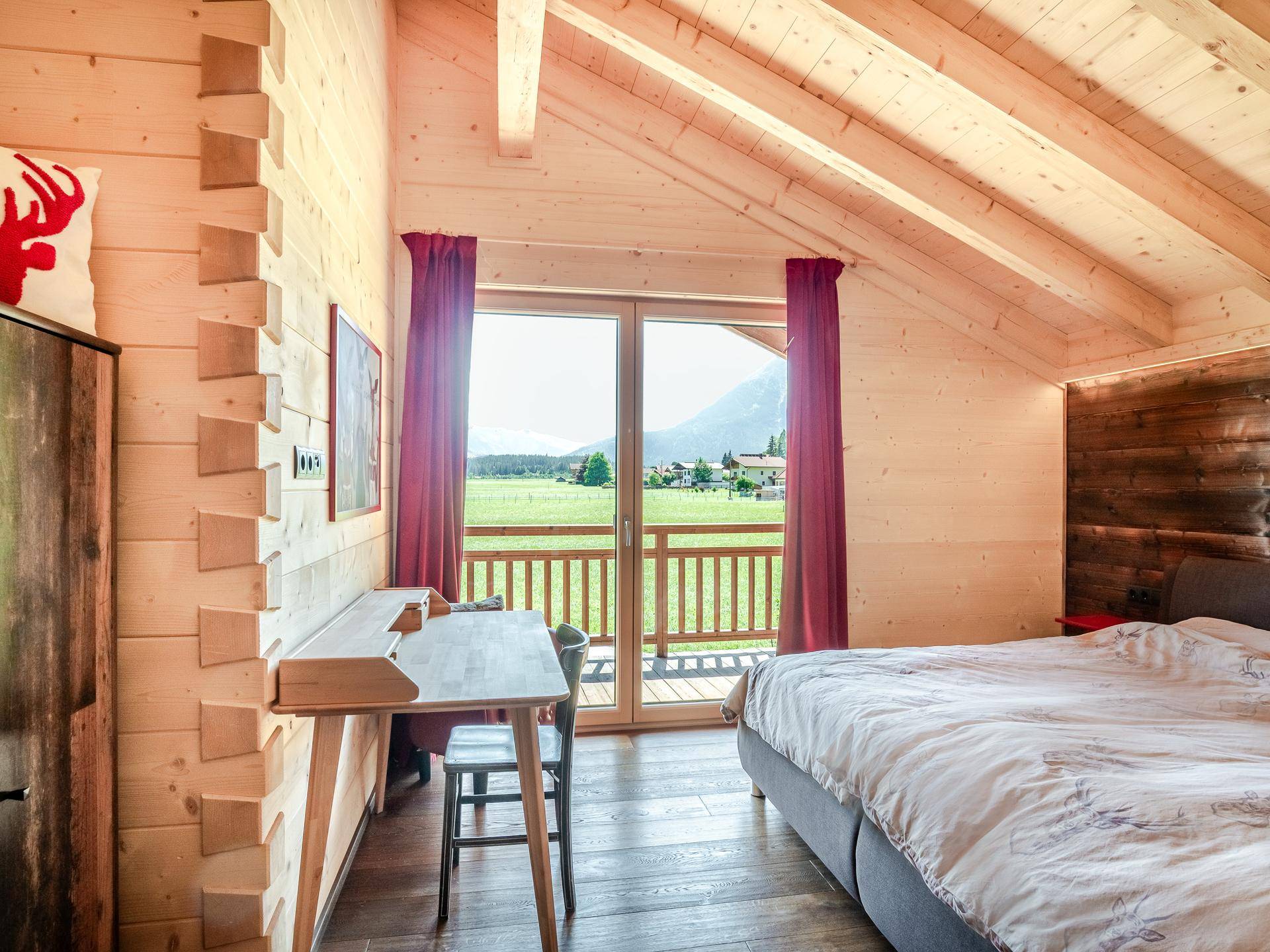 Geräumiges Schlafzimmer mit Holzbalkendecke, Schreibtisch und Balkontür, die einen weiten Blick ins Grüne bietet.
