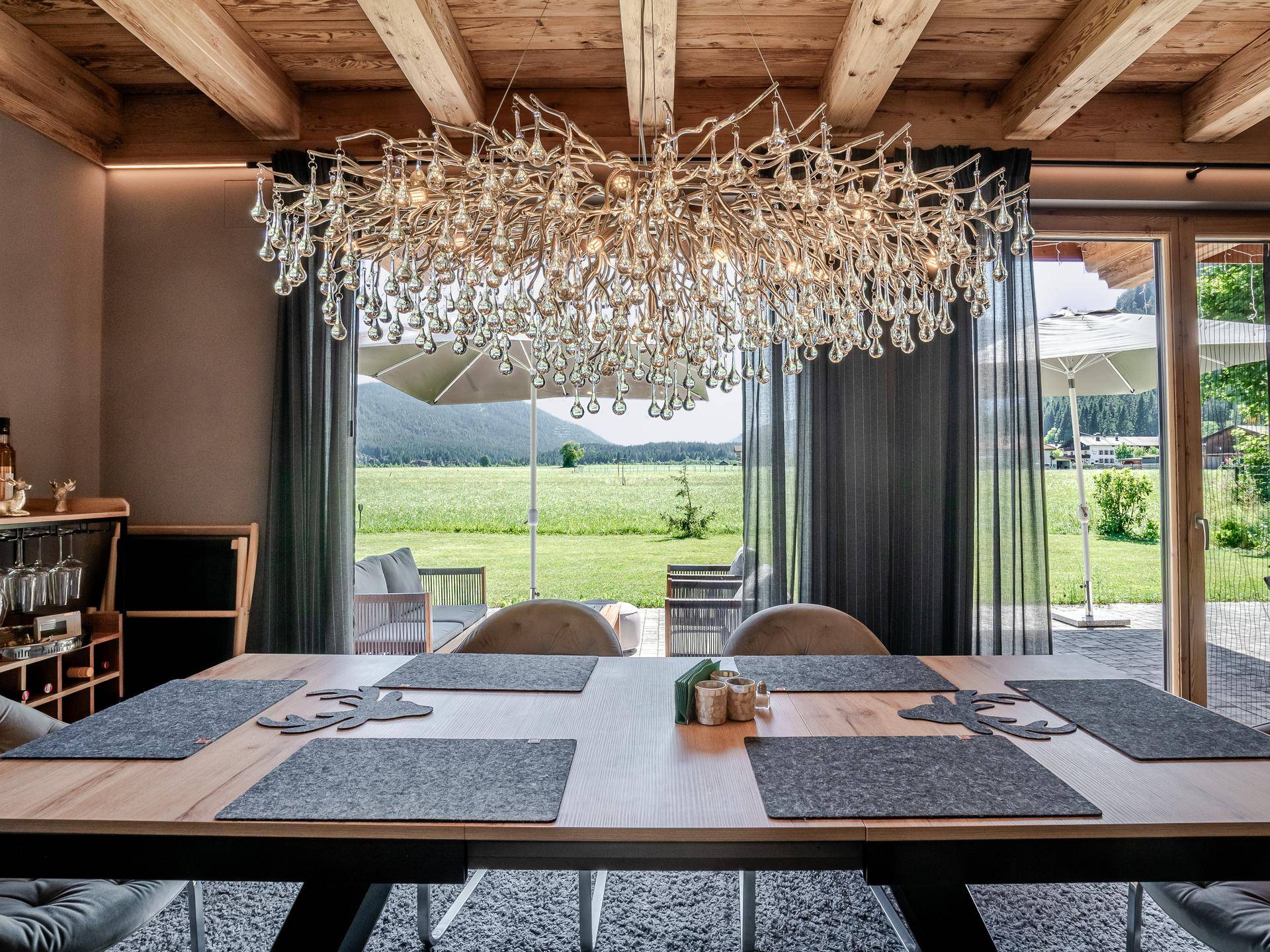Eleganter Essbereich mit Holzbalkendecke, modernem Kronleuchter und großen Fenstern mit Blick ins Grüne.