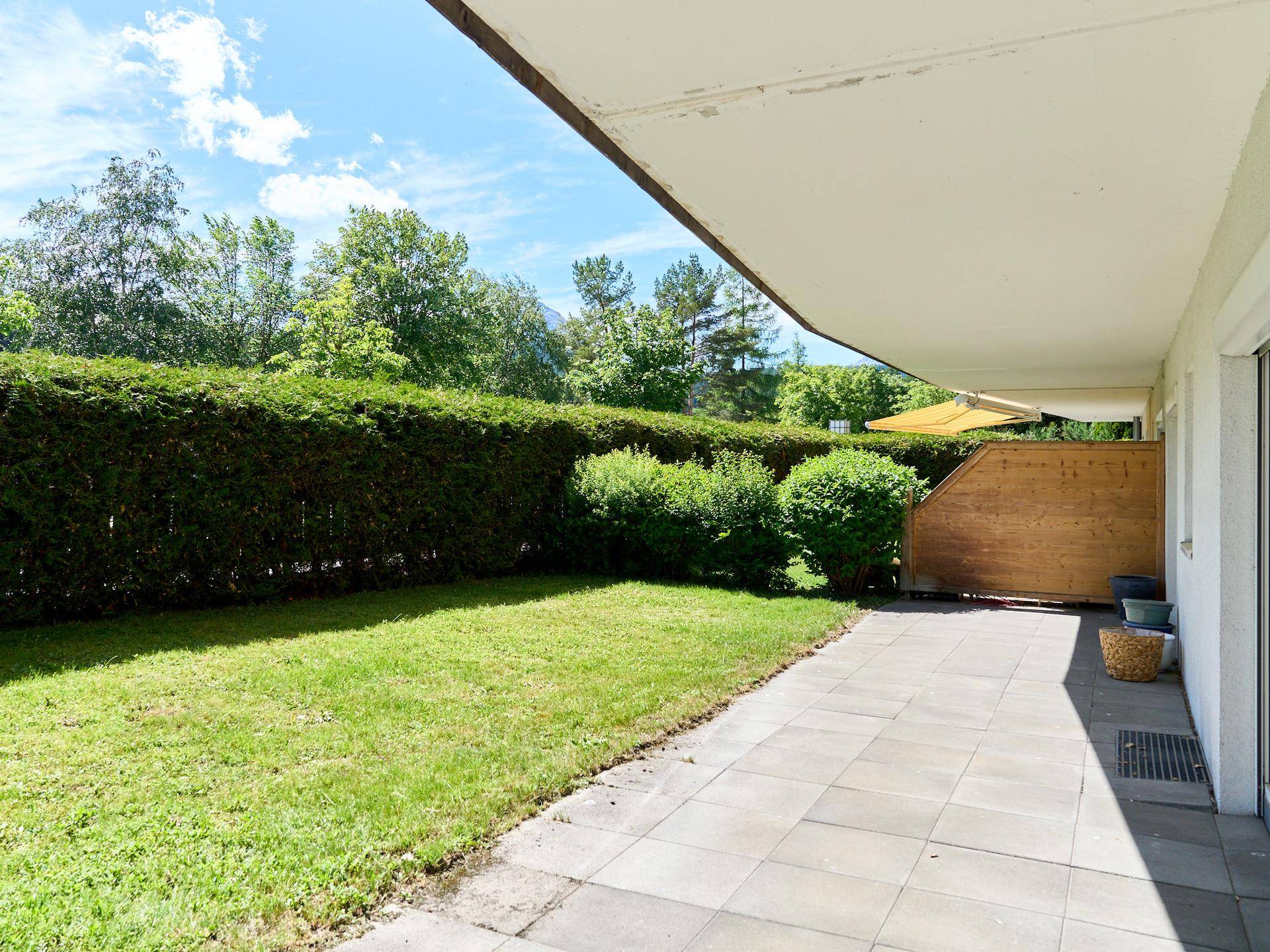 Gepflegter Gartenbereich mit Rasenfläche und gepflasterter Terrasse, umgeben von einer Hecke.